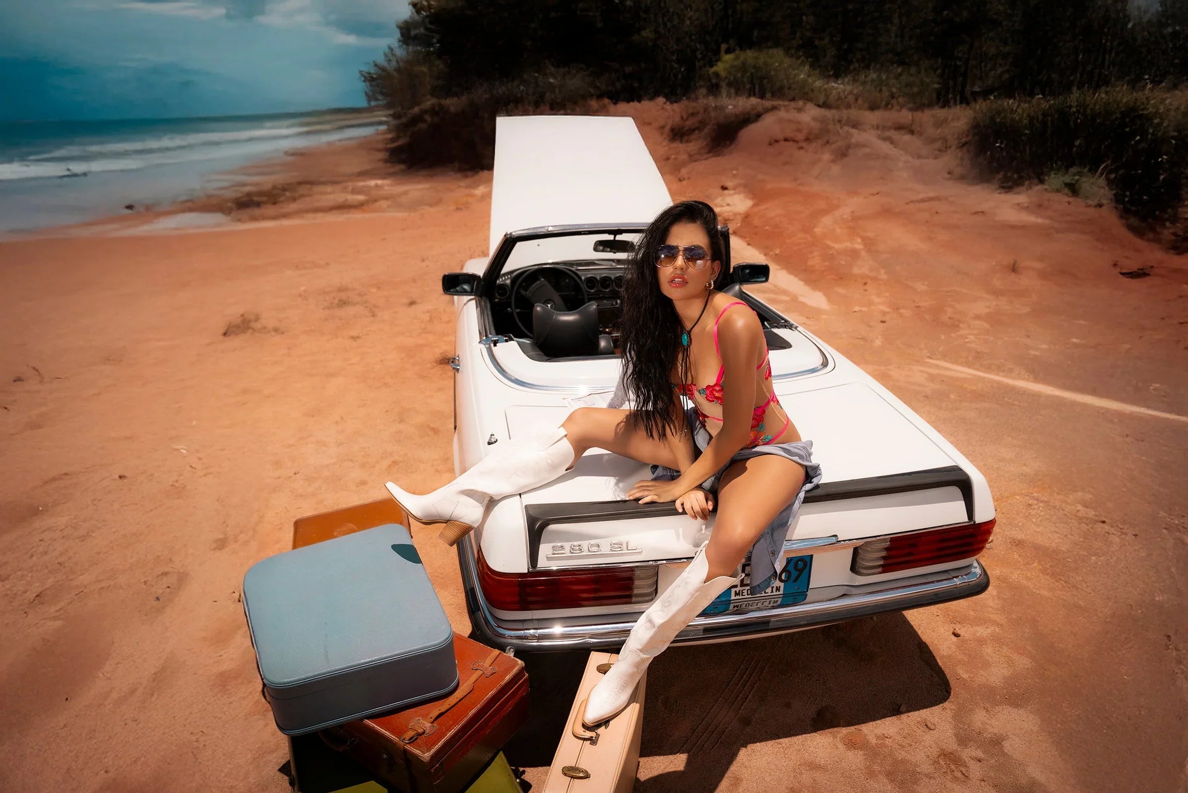 Model in Sheer Mesh Bodysuit Mapale Wild Flower posing on a beach with vintage convertible and suitcases, styled with white boots.
