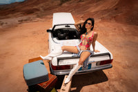 Model sitting on convertible trunk in Sheer Mesh Bodysuit Mapale Wild Flower with white boots, surrounded by luggage in desert scene.